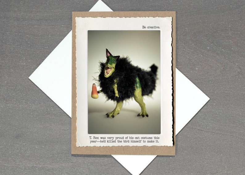 May include: Greeting card featuring a T-Rex dinosaur in a cat costume, holding candy corn. The card has the text "Be creative" and a humorous caption about the dinosaur's costume. The card is on a gray surface with a white envelope.