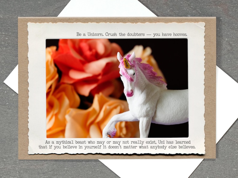 May include: A greeting card featuring a white unicorn figurine with a pink mane and horn, set against a backdrop of orange and yellow flowers. The card includes the text: "Be a Unicorn. Crush the doubters - you have hooves."