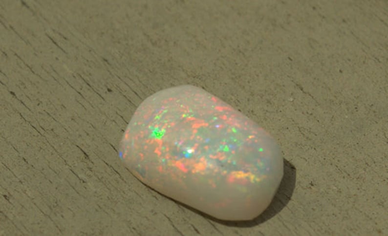 May include: A white opal gemstone with iridescent rainbow colors. The opal is a smooth, oval shape and has a natural, organic look.
