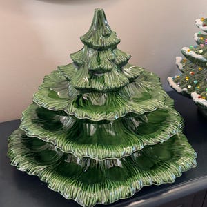 May include: A three-tiered, dark green ceramic serving dish shaped like a pine tree. The dish features a glossy finish with molded, textured boughs that create three distinct levels for serving food, with a pointed top section as the lid.