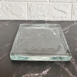 May include: A clear, square glass coaster with rounded edges. The coaster is transparent, allowing the surface beneath to be visible. The glass has a slightly textured appearance. The coaster is designed to protect surfaces from drink condensation.