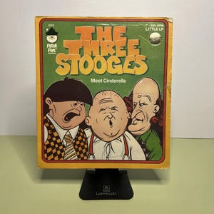 May include: Vintage "The Three Stooges Meet Cinderella" LP record album cover. The cover features cartoon illustrations of the Three Stooges with text in orange and white on a green background. The album is supported by a black stand.