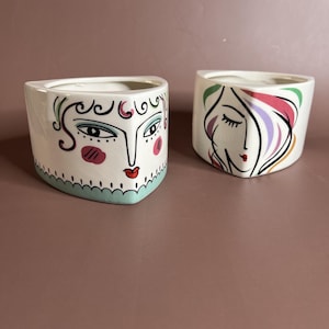 May include: Two white ceramic planters with abstract face designs. One features a face with black outlines, blue eyes, and pink blush. The other has a face with closed eyes and colorful hair. Both have a triangular shape.