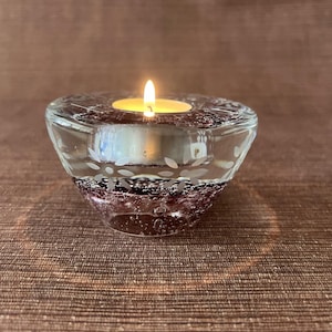 May include: A clear glass votive candle holder with a lit tea light. The holder has a decorative leaf pattern etched around the rim and a dark purple base with bubbles. The candle flame is bright yellow.
