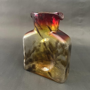 May include: A decorative glass bottle with a square base and a flared, wavy top. The bottle transitions from a deep red at the top to a golden-brown hue towards the base. The glass has a textured, swirling design.