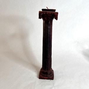 May include: A dark red pillar candle with a fluted column design. The candle is shaped like a classical column, with a square base and a decorative top. The candle has a single wick and is made of wax.