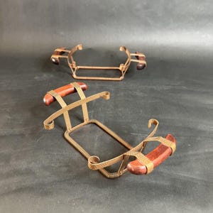 May include: Two decorative metal wine bottle holders. Each holder has a rectangular base, a woven metal frame, and wooden handles. The holders are made of copper-colored metal and dark wood. The background is a dark gray.