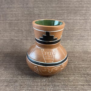 May include: A brown ceramic vase with a rounded body and a narrow neck. The vase features black and white geometric designs and a green interior. The vase is likely handmade and has a rustic, artisanal feel.