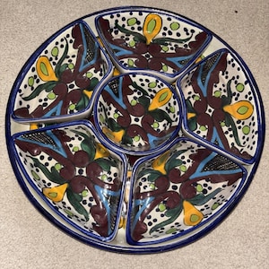 May include: A round, divided serving dish with a blue rim and a central bowl. The dish features five compartments with a floral design in burgundy, blue, yellow, and green on a white background. The design includes dots and stylized floral elements.