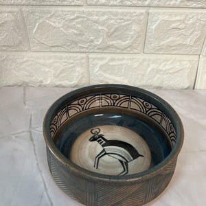 May include: A round ceramic bowl with a dark blue exterior and a cream-colored interior. The bowl features a black and white antelope design in the center and geometric patterns around the rim. The exterior has a textured pattern.