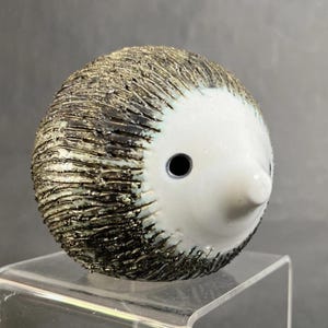 May include: A ceramic hedgehog figurine with a white face and a body covered in textured, spiky, dark brown and gold accents. The hedgehog has a small, black eye and a pointed nose. The figurine is displayed on a clear, rectangular stand.