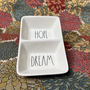 May include: A white ceramic divided dish with the words "HOPE" and "DREAM" in black handwritten font. The dish has a rectangular shape with rounded corners and is sitting on a floral patterned surface.