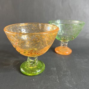 May include: Two colorful, hand-blown glass dessert cups. One is orange with green and red speckles, the other is green with white speckles. Both have a twisted stem and a round base. The cups are on a dark gray surface.