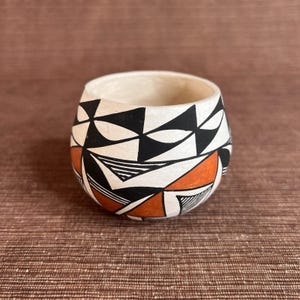 May include: A small, round ceramic bowl with a cream-colored interior and a geometric design. The exterior features black, white, and orange triangular and linear patterns. The bowl is likely handmade and has a rustic aesthetic.