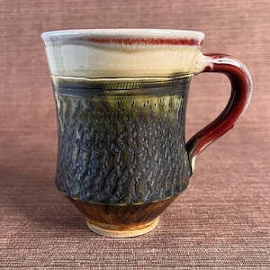 May include: A handmade ceramic mug with a textured, dark blue and green exterior. The top rim is white with a red accent, and the handle is a deep red. The mug has a unique, artisanal appearance.