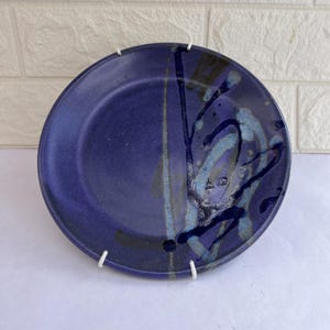May include: A round, ceramic plate with a deep blue glaze. The plate features abstract brushstrokes in shades of blue, gray, and black. The plate is displayed on a white stand against a white and brick background.