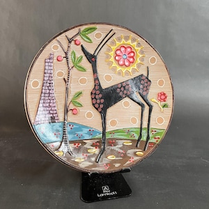 May include: A decorative ceramic plate featuring a black deer with red accents, a tree with red berries, and a stylized sun. The plate has a brown rim and a beige background with circular patterns. The plate is displayed on a black stand.