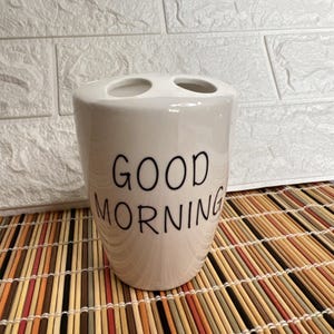May include: A white ceramic toothbrush holder with two openings at the top. The words "GOOD MORNING" are printed in black on the front. The holder is on a bamboo mat, with a white brick wall in the background.
