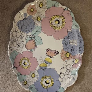 May include: Oval ceramic serving platter with a scalloped edge. The platter features a floral design with pink, blue, and yellow flowers, along with a butterfly. The flowers are arranged around the edge of the platter.