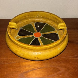 May include: A round, yellow ashtray with a decorative interior. The interior features a floral design with black, green, and orange segments. The ashtray has two built-in rests for cigarettes.