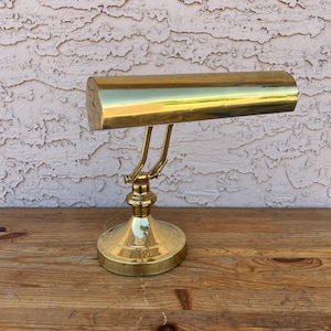 May include: A vintage brass desk lamp with a cylindrical shade and a curved arm. The lamp has a round base and a polished finish, reflecting the light. The lamp is designed to sit on a desk or table.