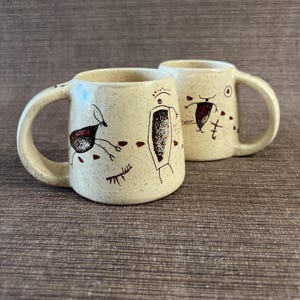 May include: Two cream-colored ceramic mugs with a speckled finish, each featuring dark brown hand-drawn petroglyph-style figures and abstract markings. The mugs have rounded bodies and curved handles, showcasing a consistent artistic design.
