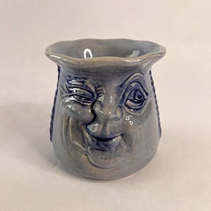 May include: A blue ceramic mug with a winking face design. The mug features a smiling face with detailed facial features, including a winking eye, nose, and mouth. Braided details are on the sides of the mug.