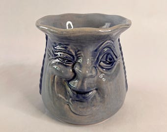 May include: A blue ceramic mug with a winking face design. The mug features a smiling face with detailed facial features, including a winking eye, nose, and mouth. Braided details are on the sides of the mug.
