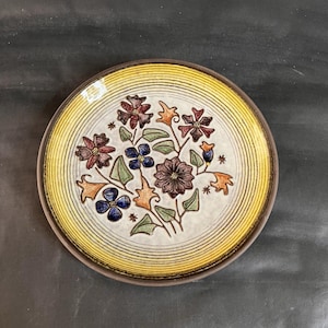 May include: A round ceramic plate with a floral design. The plate features a cream-colored center with a raised floral pattern in shades of brown, blue, orange, and green. The rim is yellow with concentric lines, and the outer edge is brown.
