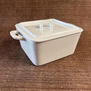 May include: A white, square ceramic casserole dish with a matching lid. The dish has a handle on each side and a loop handle on the lid. The dish is sitting on a brown surface.