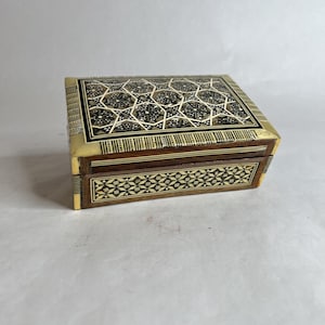 May include: A rectangular wooden box with intricate inlaid geometric patterns. The lid features a repeating star and floral design in shades of black, white, and brown. The box has brass corner accents and a decorative border.