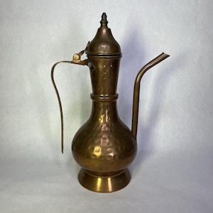 May include: A vintage copper pitcher with a long spout and a curved handle. The pitcher has a bulbous body and a lid with a small finial. The copper has a hammered texture and a warm, antique finish.