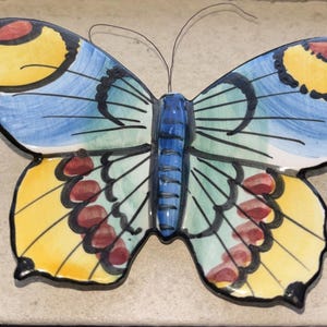 May include: A ceramic butterfly with a colorful design. The wings are painted with yellow, blue, and green hues, accented with red and black details. The butterfly has a blue body and black antennae.