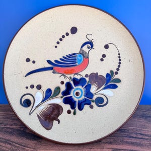 May include: A ceramic plate with a hand-painted design of a blue and red bird perched on a blue flower with brown and green leaves. The plate has a cream background with brown accents.