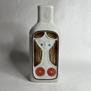May include: White ceramic bottle-shaped vase with a textured surface. The vase features a stylized woman's face with blue eyes, brown hair, and two orange circles. The design is framed by a dark brown border.