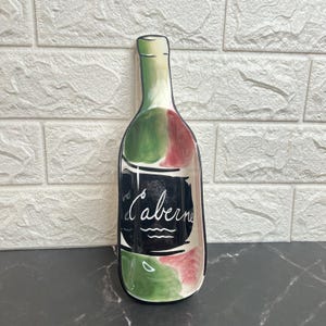 May include: A ceramic wine bottle-shaped serving dish. The dish is painted with green, red, and white accents. A black label on the front reads "Cabernet" in white script. The dish is on a black surface.