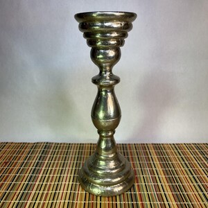 May include: A metallic silver candlestick holder with a tiered design. The base is wide and circular, leading to a slender, ornate stem. The top has a wide, shallow cup for holding a candle. The candlestick is on a striped surface.