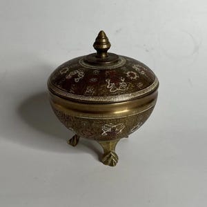 May include: A small, antique brass container with a lid and three decorative feet. The container is adorned with floral patterns in red and white. The lid is topped with a small, pointed finial. The container is likely used for storing small items.