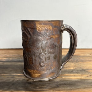 May include: A handcrafted ceramic mug with a dark brown and gold metallic glaze. The mug features embossed details and a curved handle. The mug sits on a wooden surface.