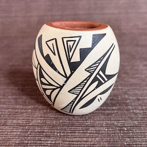 May include: A small, round, cream-colored ceramic pot with a reddish-brown rim. The pot is decorated with black geometric patterns, including triangles, lines, and abstract shapes. The design covers the majority of the pot's surface.