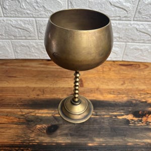 May include: A vintage brass goblet with a round bowl and a beaded stem. The goblet sits on a circular base. The brass has a warm, aged patina. The background is a wooden surface and a white brick wall.