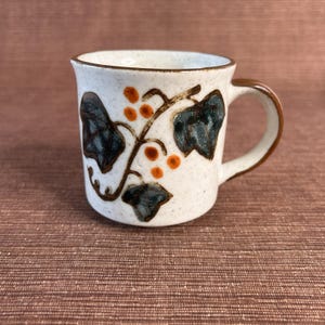 May include: A ceramic coffee mug with a cream-colored speckled finish. The mug features a brown handle and rim, and is decorated with a vine design with dark green leaves and orange berries. The mug is a standard size for hot beverages.