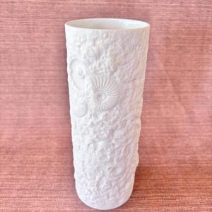 May include: A white ceramic vase with a textured surface and embossed fossil designs. The cylindrical vase has a smooth rim and a slightly rounded base. The vase is approximately 8 inches tall.