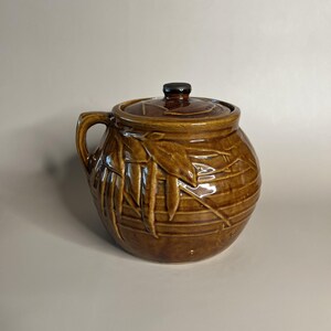 May include: A brown ceramic jar with a lid and handle. The jar has a glossy finish and is decorated with a leaf pattern. The lid has a small, dark knob. The jar is likely used for storage or display.
