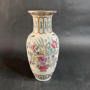 May include: A cream-colored ceramic vase with a floral design. The vase features a red base, a bouquet of pink, yellow, and purple flowers, and green leaves. The rim and neck are decorated with gold and green accents. The vase is approximately 10 inches tall.