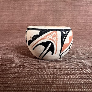 May include: A small, round, hand-painted ceramic bowl. The bowl features a cream-colored base with black and coral-colored geometric designs. The rim is painted black, and the bowl has a smooth, rounded shape.
