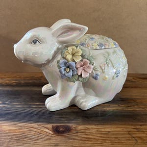 May include: A ceramic bunny-shaped trinket box with a pearlescent finish. The bunny is decorated with pastel-colored flowers and has a lid on its back. The box is sitting on a wooden surface.