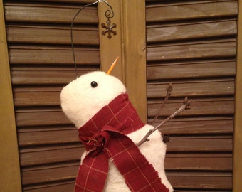 May include: A handmade snowman ornament with a red tartan scarf, twig arms, and a carrot nose. The snowman has a black eye and a snowflake decoration. It rests on a wooden base with greenery and red berries.
