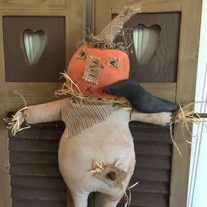 May include: A handmade scarecrow doll with an orange pumpkin head, stitched patches, and a burlap hat. The scarecrow has a striped shirt, straw arms, and legs. It is a rustic, fall-themed decoration.