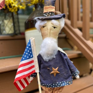 May include: A handcrafted Uncle Sam doll, dressed in a blue coat with a star, striped pants, and a top hat. He holds a small American flag. The doll is standing on a wooden base, perfect for patriotic decor.